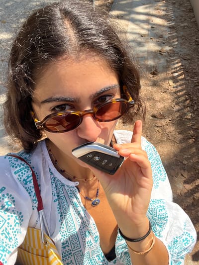 A young woman, partially visible, is depicted in a close-up, high-angle self-portrait outdoors in Caminha, Portugal. She has dark, short to medium-length hair with a central part and wears tortoiseshell oval sunglasses with brown lenses. Her dark eyes are visible above the sunglasses, looking directly at the viewer. She is wearing a white blouse with a teal geometric or abstract pattern. Around her neck are two necklaces: a shorter gold-colored chain with small red beads and a longer gold-colored chain featuring blue beads and a central oval blue pendant with a gold-colored frame. On her right wrist, a black elastic band and a thin gold-colored bangle are present. Her right hand holds a black and silver BMW car key, with the BMW logo visible, positioned near her mouth. A yellow and white striped bag with a red strap is partially visible on her left side. The background consists of a dry, unpaved ground surface with scattered dry leaves and debris, illuminated by strong direct sunlight creating distinct shadows.
