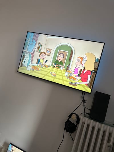The image displays an indoor residential setting in Gouvieux, France, primarily featuring a large, wall-mounted flat-screen television. The television screen shows an animated scene from "Rick and Morty," depicting five characters – Rick, Morty, Jerry, Summer, and Beth – seated around a dining table, eating pancakes. Rick, on the far left, holds a fork. Morty, next to Rick, is looking at a smartphone. Jerry is visible through an archway in the background, holding cutlery. Summer and Beth are on the right side of the table, also holding cutlery. The animated table is set with multiple plates of pancakes, mugs, a stick of butter, and small glasses of orange juice. Two framed pictures, one of a horse and one of a butterfly, adorn the wall within the animated scene. Blue backlighting emanates from behind the television. Below the television, a black console (possibly a gaming system) is positioned on a transparent surface, above a white panel radiator. A black wired gaming headset with circular earcups hangs on the wall to the left of the console. Several black cables are visible, connecting various electronic components. The walls are a plain, light color. A partial view of a second screen displaying animated content is visible in the lower-left corner of the image.