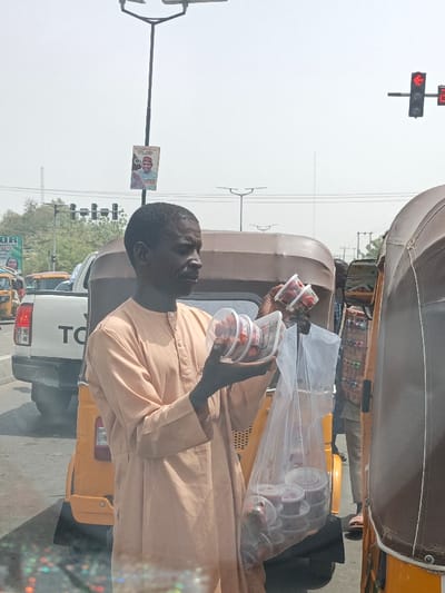 An adult male stands on a street in Hotoro, Nigeria, displaying items for sale. He possesses dark skin and short dark hair, wearing a light peach-colored long-sleeved tunic with a buttoned placket. In his hands, he holds multiple clear plastic containers, each filled with red strawberries. A larger clear plastic bag, also containing several identical strawberry containers, hangs from his left hand. He is positioned between stopped vehicles, with a yellow three-wheeled auto-rickshaw behind him and the rear of a white Toyota pickup truck visible further back. To his right, the front portion of a brown vehicle is visible. A street light pole with solar panels rises in the background, featuring a political poster depicting a man in a green cap with the text "MUNKUDALLAH." A traffic light showing a red signal is visible to the far right. The sky is hazy and bright.