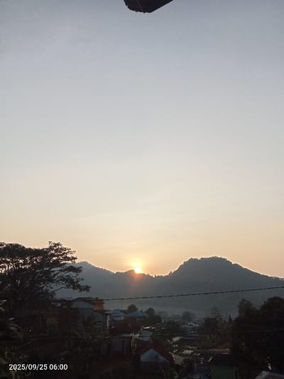 The image displays a sunrise over a residential area in Ciputri, Indonesia, documented on 2025/09/25 at 06:00. The sun, a bright orange orb, is visible at the crest of a dark, forested mountain range that forms the horizon line. The sky above transitions from a pale yellow-orange near the sun to a light grey overhead, encompassing the upper two-thirds of the frame. A dark, indistinct structure overhangs the top center. Below the mountains, a collection of residential structures with various roof colors (e.g., red, blue) is partially visible amidst dense, dark green foliage and trees. A large, dark-canopied tree occupies the left foreground, silhouetted against the rising light. Multiple dark, thin horizontal lines, consistent with utility wires, intersect the lower mid-ground.