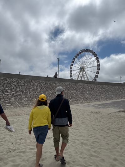 Two individuals are walking on a sandy beach, viewed from behind. The person on the left is wearing a yellow hooded sweatshirt and blue denim shorts, with a yellow baseball cap. The person on the right is wearing a gray baseball cap, a dark jacket with light gray accents, and khaki shorts. The beach is composed of light-colored sand. In the background, there is a stone wall and a promenade. Beyond the promenade, a large Ferris wheel is visible against a cloudy sky with patches of blue. Several birds are flying in the sky. A person is carrying a white sneaker in their left hand. This scene is located in Berck, France, likely near the waterfront and Ferris wheel.