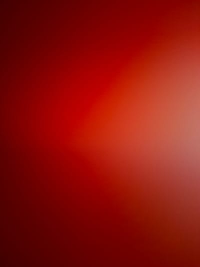 This is an abstract image featuring a gradient of red and orange colors. The image is blurred, with a bright light source creating a luminous area on the right side. The dominant color is a deep red that transitions to lighter shades of orange and a bright whiteish-yellow where the light is most intense. There are no discernable objects, people, or environmental features visible. The image is a pure color gradient with no specific location or context that can be identified.