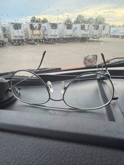 An overhead, close-up shot captures a pair of thin-framed glasses resting on a black, textured surface, possibly a dashboard, inside a vehicle. The glasses have clear, round lenses and thin silver frames. The left lens appears slightly detached or bent upwards at the frame. In the background, viewed through a rain-streaked window, a line of white refrigerated semi-trailers are parked in a lot under an overcast sky with hints of sunlight. The scene suggests a roadside stop or parking area, common in commercial trucking hubs. The weather appears to be rainy, with water droplets visible on the window. No people are visible in the image.