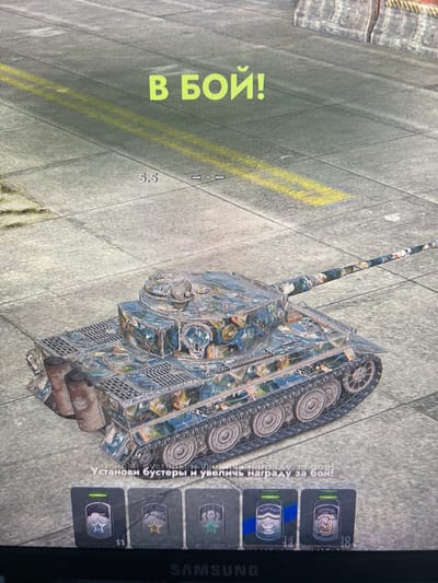 The image shows a screenshot from a video game. The foreground features a camouflaged tank, likely a representation of a World War II-era German Tiger tank, on a paved surface with yellow lines. In the background, there appears to be a city or industrial setting with buildings and structures. Above the tank, the text "В БОЙ!" is displayed in large yellow letters, which translates to "Into battle!" from Russian. Below the tank, there is text in Russian that reads "Установи бустеры и увеличь награду за бой!", meaning "Install boosters and increase your battle reward!". There are also icons representing in-game items at the bottom of the screen. The scene does not depict any specific location in Bely Klyuch, Russia, as it is a digital representation within a game.