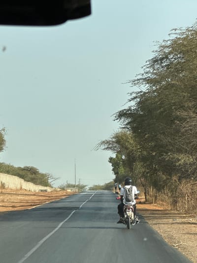 The image shows a motorcycle traveling on a paved road in the Communauté rurale de Malicounda, Senegal. The rider wears a white shirt, dark pants, and a helmet. The road has a white line marking. There are trees and vegetation on both sides of the road. The sky is clear and light blue. A portion of a vehicle is visible in the lower left corner.