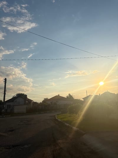 The image depicts a street in Kukorevo, Bulgaria, taken during a bright, sunny day. The sun is high in the sky, casting strong light and shadows. Power lines stretch across the frame. Houses are visible along the side of the road, with vegetation and trees interspersed. The road itself appears to be paved but somewhat worn. There are no people visible in the shot. The overall impression is that of a quiet, rural or suburban street on a clear day.