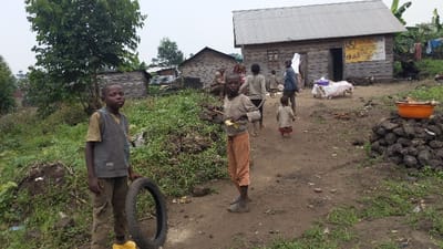 A rural scene depicts children in worn clothing, standing in a dirt path surrounded by lush green vegetation and simple wooden houses. One child holds a tire. Several other people are visible in the background, some walking. An orange bowl sits atop a pile of rocks near a building with a poster on its exterior. The overall atmosphere is one of poverty and simplicity.