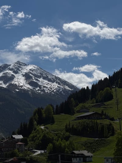 The image shows a mountain landscape in Bad Gastein, Austria. The left side of the frame is dominated by a mountain covered in snow patches under a blue sky with scattered white clouds. A dense forest covers the lower slopes of the mountain, transitioning into a green hillside with buildings and a ski lift at the top right. Some buildings and a road are visible in the bottom right corner, indicating a town.