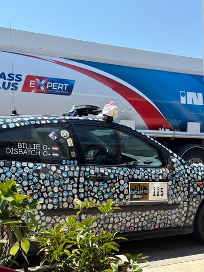 This image shows the side of a dark-colored car heavily adorned with numerous small stickers, many of which appear to be related to technology and programming. A larger sticker on the driver's side door displays "115" and "2026 Garbage run Albania," suggesting the car might be participating in a rally or event. Adjacent to this, text reads "BILLIE O+ DISBATCH O+" with two small flag stickers, one of which is the Dutch flag.

In the background, a large white truck with blue and red stripes is partially visible. The truck has "IN" and "EXPERT FORMULA" text on its side. The scene appears to be outdoors during the daytime, with clear blue sky visible on the left. Some green foliage is present in the foreground on the right, partially obscuring the car and truck. The overall impression is of a custom-decorated vehicle, possibly at a stop during a road trip or event. No people are clearly visible in the frame. The lighting suggests it is a sunny day.