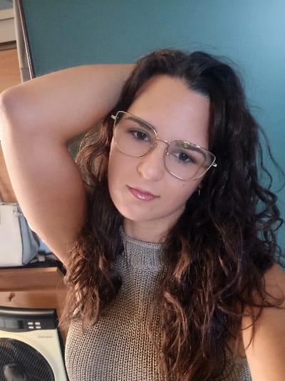 The specific location within San Marzano sul Sarno, Italy, cannot be determined from the visual information provided in the image. The scene is an indoor, close-up shot of a single female subject.
The subject is a light-skinned female with long, wavy, dark brown hair. She wears gold-framed, cat-eye eyeglasses and a light gray, ribbed knit, sleeveless top with a high neck. Her lips are glossy and appear to be covered in a light pink or nude shade. She is posed with her right arm raised, with her hand positioned behind her head, mostly out of frame. Her eyes are open, and her gaze is directed forward.
The background features a solid teal-colored wall behind the subject. To the lower left, a portion of light-colored wooden furniture is visible, on which a white and light blue handbag is placed. Below the furniture, a cream-colored electric fan with a black grille is partially visible.