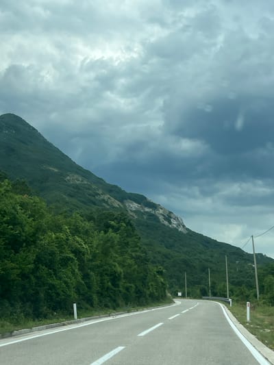 The image shows a winding asphalt road in the city of Dragalj, Montenegro. The road is bordered by white lines and has a concrete curb. To the left of the road, a steep, green mountain rises, covered in dense foliage. The upper part of the mountain reveals rocky, lighter-colored patches. On the right side of the road, a few utility poles with wires are visible, and the terrain is grassy with some scattered vegetation. The sky is overcast with dramatic, dark clouds, suggesting an impending storm. There are no people or vehicles visible in the scene.