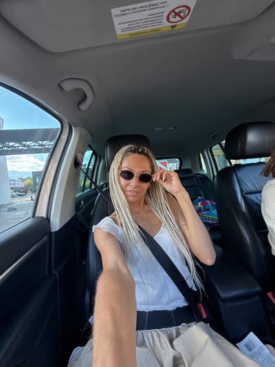 A woman with blonde braided hair and sunglasses is sitting in the driver's seat of a car. She is wearing a white top and a beige skirt, and her seatbelt is fastened. The interior of the car is visible, including the seats, dashboard, and a portion of the roof. The background shows a street scene with some buildings and a cloudy sky. The location is identified as Есбол ауылдық округі, Kazakhstan.