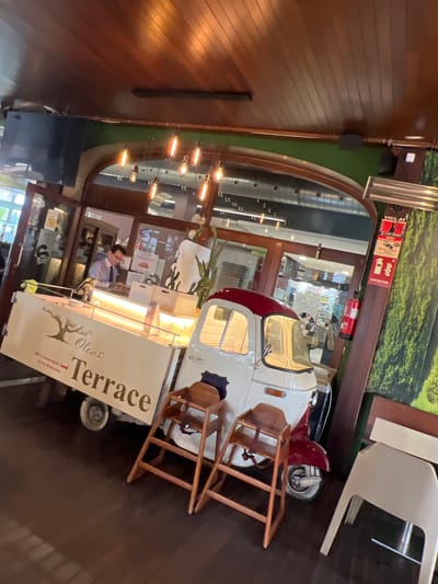 The image shows the interior of a restaurant or cafe. A wooden-paneled wall is on the left, and a decorative archway leads to another area. Inside the archway, there is a food stall or service counter resembling a vintage vehicle, with the text "Olens Terrace" on its side. A person is behind the counter, likely a server or employee. Further back, there's a glimpse into what seems like a shop or display area. The location is likely Cambrils, Spain, inferred from the text on the stall referencing "Olens Terrace" and potentially related to local businesses or features of Cambrils. A chair is visible near the stall, suggesting seating for customers.