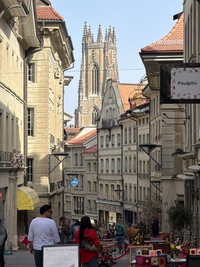 The image depicts a bustling street scene in Fribourg - Freiburg, Switzerland, specifically looking north-east along Rue de Lausanne, with the tower of St. Nicholas Cathedral visible in the background.

The environment is characterized by densely packed historic multi-story buildings constructed primarily of light-colored stone, featuring numerous windows, balconies, and red-tiled pitched roofs. The street slopes gently downward. In the distant background, the tall, ornate Gothic tower of St. Nicholas Cathedral dominates the skyline, exhibiting intricate spires, arched windows, and detailed stonework under a clear blue sky.

Along the street, commercial activity is evident. On the right, a dark sign with "Poulpito" in white text and stylized marine life hangs above the street. Further down, another dark sign with gold lettering displays "Bindella." On the left side of the street, a blue circular sign with "DURING" in white text is mounted on a building.

In the midground, multiple pedestrians are visible, moving along the street. Outdoor seating areas with tables and chairs are set up on the right side, defined by a red rope barrier. Adjacent to these tables, a display features colorful knitted or crocheted squares, possibly textiles, on a wooden surface, alongside buckets containing flowers. A small leafy tree is planted nearby.

In the foreground, two individuals are prominent, walking away from the camera. The person on the left wears a white short-sleeved top and dark trousers. The person on the right wears a red long-sleeved top, dark trousers, and carries a light-brown shoulder bag. Positioned between these two individuals, closer to the viewer, is a black A-frame sign with white text advertising "Restaurant Galata," listing "CARTE" (menu) details. On the immediate left, a building facade features a yellow ribbed awning and ornate stone carvings, including cherub-like figures, on a balcony railing. The scene is brightly illuminated by direct sunlight.