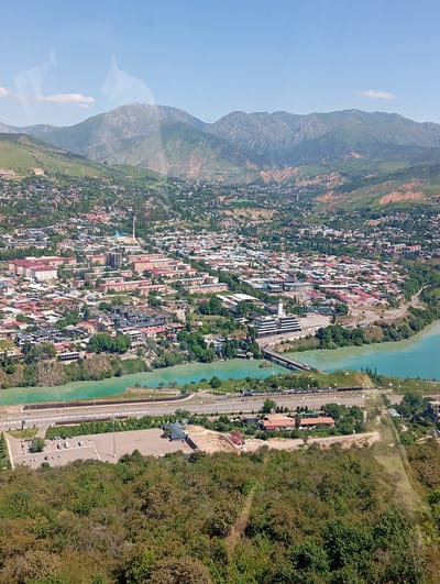 The image shows a high-angle view of Charvak, Uzbekistan, encompassing a cityscape nestled in a valley surrounded by mountains. A river of turquoise water meanders through the city, crossed by a bridge. The city is a mix of residential and commercial buildings, with various architectural styles visible. Green trees and vegetation are interspersed throughout the urban landscape. At the base of the frame, vegetation covers the foreground, with a parking lot and highway visible nearby. The sky is clear with a few scattered clouds.