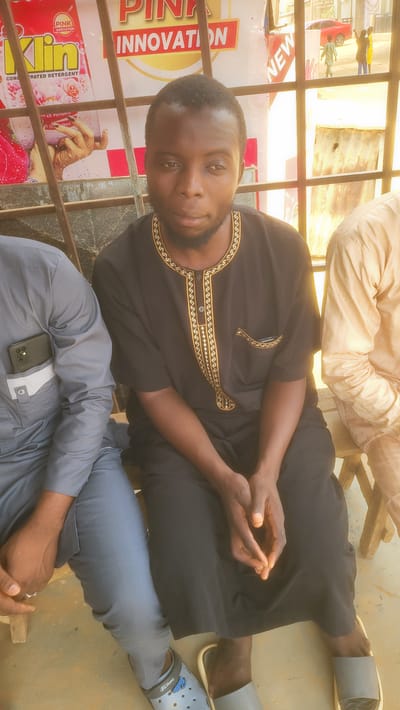 A man is seated in an outdoor setting. He has dark skin, short hair, and a beard, and he is wearing a black shirt with gold embroidered patterns around the neckline and a pocket. His hands are clasped together in his lap. To his right, another individual is wearing a light brown top, and to his left, another person is wearing gray attire. The man is wearing gray rubber slippers on his feet. Behind the man, there's a sign that says "PINK INNOVATION" and has images of products. It appears to be near a window frame or open area, with other buildings visible in the background. The location may be in Jogana, Nigeria, based on clues from the signage and appearance of the individuals.
