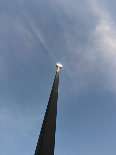 A street lamp stands tall against a pale blue sky with wispy clouds. The lamp is square and emits a bright white light, appearing to be on. The pole of the lamp is dark gray and tapers to a point as it extends upwards. There are no people or other objects visible in the frame. The image is taken from a low angle, looking up towards the sky. The location in the city of Cardwell, United States is unknown.