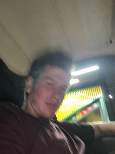 The media file is a low-resolution, motion-blurred image of a person lying down. The person appears to be a young male with short, dark hair and fair skin. He is wearing a dark-colored top. His face is partially obscured and his expression is difficult to discern due to the blur.
The background is dark and features bright, vertical lights, suggesting an indoor artificial environment. To the right, a blurred object with green and yellow bands is visible, possibly part of a machine or equipment. There is no clear indication of the time of day, weather, or specific location beyond the provided context of Lenin, Kyrgyzstan. The overall impression is one of a candid, possibly spontaneous, capture, given the quality and composition. The motion blur suggests movement during the photo capture.