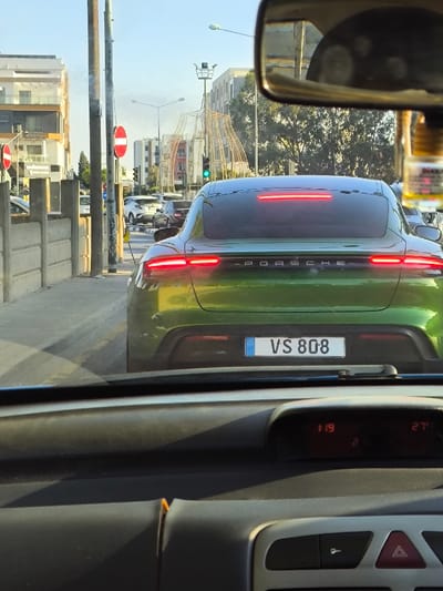 The image presents a forward-facing view from inside a stationary vehicle, capturing a street scene in Gonyeli, Northern Cyprus. In the immediate foreground, the observer's vehicle dashboard is visible, featuring a digital display showing "119" and "27" (likely time and temperature) and a hazard light button. A small, amber-colored hanging air freshener or charm dangles from the rearview mirror, which partially reflects an interior ceiling.

The central subject is a metallic green, possibly chameleon-painted, Porsche Taycan sedan directly in front, displaying a Northern Cyprus license plate "VS 808". Its full-width rear LED taillight strip and lower brake lights are illuminated red, indicating it is stopped or braking. The word "PORSCHE" is emblazoned across its rear.

To the left of the road, a sidewalk is bordered by low, grey concrete barriers. A utility pole stands on the sidewalk, bearing a red circular 'No Entry' sign, with another identical sign visible further down the street. Overhead wires are suspended from the poles. In the mid-ground, a green traffic light is illuminated for the observer's direction of travel.

The background features multiple modern, multi-story buildings, typical of urban development, lining both sides of the street. A distinctive large, abstract, skeletal metal structure, resembling an archway or decorative installation, stands prominently at a distance, identifying the location as the **Gönyeli Roundabout (Gönyeli Çemberi)** in Gonyeli. Other vehicles, including a white SUV, are visible in the traffic lane ahead of the Porsche. The sky is clear and bright, consistent with daytime conditions.