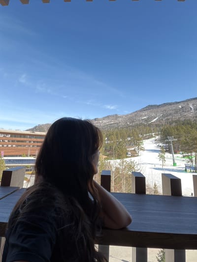 A woman sits at a wooden structure overlooking a snowy mountain and a building. The sky is blue. It is not possible to confirm the location of the image based on the visual information.