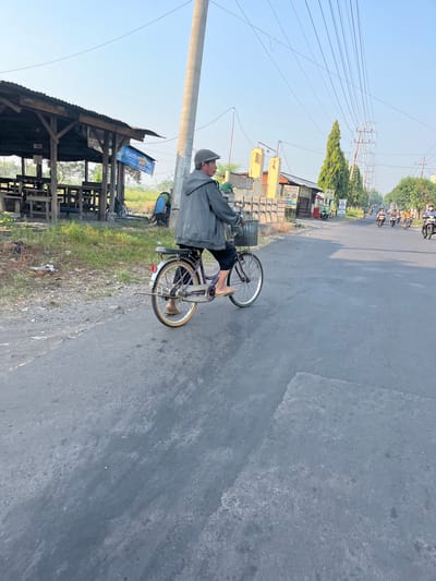 The image depicts a street scene in Sugih Waras, Indonesia, under a clear blue sky. In the foreground, a person wearing a grey jacket and a cap rides a purple bicycle with a basket on the handlebars. The person is seen from behind and to the side, with their feet on the pedals. The bicycle has a rear rack and a visible "UNITED" brand on the seat. To the left of the road is a wooden structure with a corrugated metal roof, resembling a market stall or shelter, with benches and tables. A patch of green grass and some scattered debris are visible in front of the structure. The road is paved and shows signs of wear. In the background, several motorcycles are traveling in the same direction as the cyclist. Various buildings and trees line the side of the road, and power lines stretch across the sky. The overall impression is of a typical day in a town or rural area.