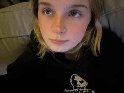 A person with blonde hair lies on a gray pillow, looking directly at the camera with slightly widened eyes. They are wearing a black hooded sweatshirt. A portion of the sweatshirt's logo is visible, featuring a graphic with the letters "SENS" above a figure of a person or animal. The lighting is dim, and the camera angle is close-up and slightly tilted.
