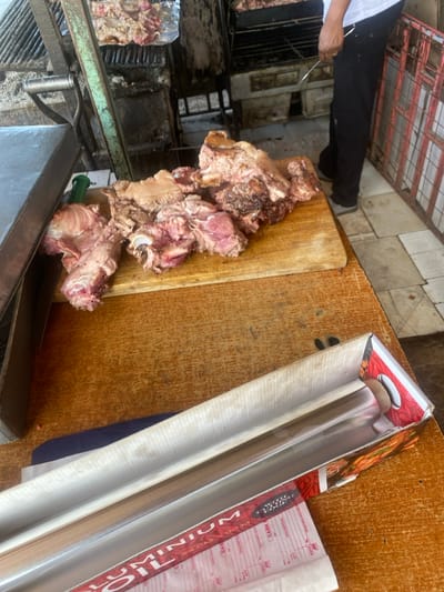 The scene depicts a food preparation area, likely a street food stall or butcher shop, in the city of Kiaga, Kenya. In the foreground, a worn wooden tabletop displays an open cardboard box containing a roll of aluminum foil. Beneath the box is a white paper form with red text. To the left, a dark gray object, possibly a scale, rests on the table. In the midground, a wooden cutting board holds several pieces of raw, butchered meat, consisting of muscle, fat, and bone, exhibiting varying shades of pink, red, and white. Directly behind this, a green metal support structure is visible. In the background, multiple metal grills are present, some containing meat actively cooking or warming, partially covered by aluminum foil. A partially visible individual, wearing a white shirt and black pants, stands near the grills, holding a long metal utensil. The floor is tiled with light-colored squares, and a metal grate or cage structure is visible to the right.