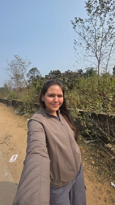A woman in a brown jacket and grey pants stands outdoors, smiling at the camera. Behind her, there's a wall with greenery, and trees frame the blue sky above. The ground is dirt or gravel, and some paper trash is visible near the bottom right corner.