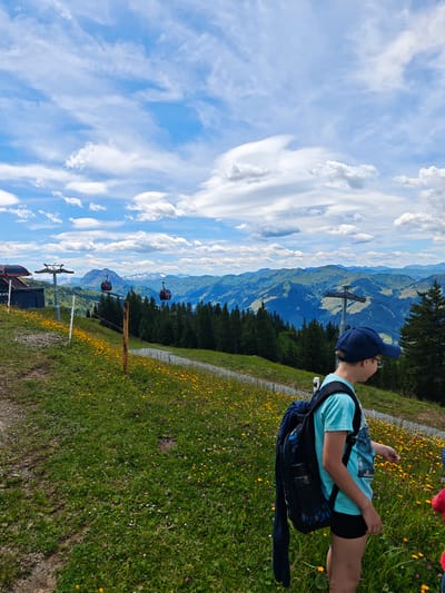 In the Austrian Alps near Kirchberg in Tirol, a young person wearing a blue baseball cap and a light blue t-shirt stands with a black backpack. They are on a grassy slope covered in yellow wildflowers, with a gravel path to their left. In the background, a ski lift with red gondolas ascends a mountain. Beyond the ski lift, layers of green mountains stretch to the horizon under a cloudy blue sky.
