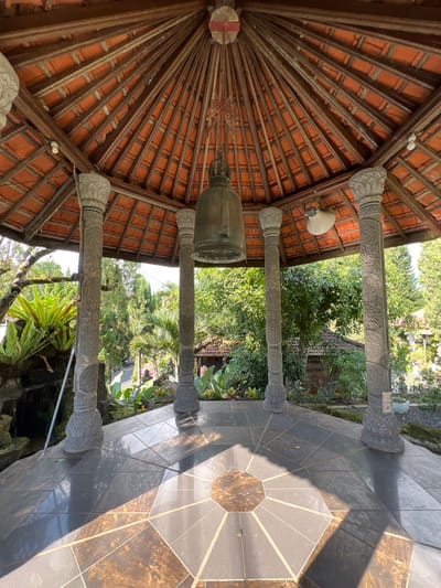 The image shows the interior of a gazebo in Pupuan, Indonesia. The gazebo has a tiled roof supported by four carved stone pillars. A large bell hangs from the center of the roof. The floor is tiled with a geometric pattern of dark and brown tiles. Lush green vegetation and trees surround the gazebo, and a small building is visible in the background. The scene is bathed in sunlight, creating strong shadows on the floor.