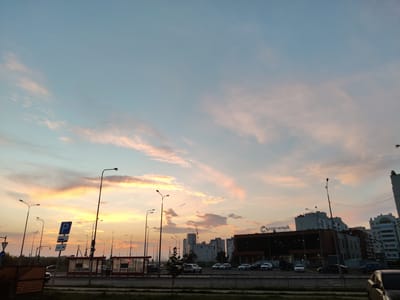The image depicts a suburban street scene at dusk in the city of Pridorozhny, Russia. The sky is filled with soft, wispy clouds tinged with hues of pink and orange from the setting sun, contrasting with the pale blue of the upper atmosphere. A row of streetlights stands tall along the side of the road, some illuminated.
In the foreground, a paved road is visible, with parked cars lining the street. A bus stop shelter with a red frame and transparent panels is situated on the right side of the road. Several vehicles are seen on the road and in the parking area. In the distance, multi-story residential buildings and a commercial building with a sign that reads "СОЛНЕЧНЫЙ" (Solnechny) are visible. The overall impression is that of a calm evening in a residential and commercial district.