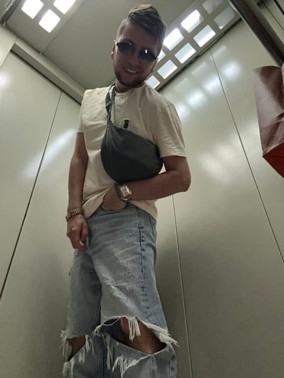 A young man with short, styled blonde hair and sunglasses poses for a selfie in an elevator. He is wearing a cream-colored t-shirt with a small black bear graphic, light blue distressed denim shorts with prominent rips, a dark gray fanny pack worn across his chest, and a gold wristwatch. He has one hand in his pocket, revealing his thumb. The elevator interior is characterized by beige-colored walls and ceiling panels, with fluorescent lighting in the ceiling. The image is shot from a low angle, looking up at the man. The scene appears to be an interior elevator car, possibly in a residential or commercial building. The provided context suggests this location is in Oktyabrskiy, Russia.