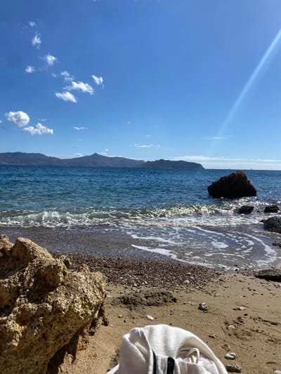 The image depicts a sunlit coastal landscape in the city of Megalochori, Greece. In the immediate foreground, a light brown sandy beach, mixed with small pebbles and darker stones, extends across the lower portion of the frame. A large, textured, light-colored rock is positioned in the lower-left. On the sand in the lower-right, a folded white garment or towel is visible. Small waves with white foam are breaking and receding at the water's edge. The mid-ground consists of clear, blue-green sea water. A distinct, dark rock formation is partially submerged in the water towards the right side. Across the sea, in the background, a range of dark mountains or hills extends along the horizon. The sky above is a vibrant blue with several scattered white cumulus clouds, primarily in the upper-left quadrant. A diagonal lens flare or sunbeam is evident in the upper-right, indicating strong direct sunlight. No individuals or specific actions, beyond the movement of waves, are discernible.