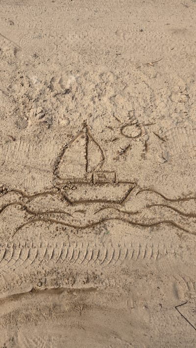 The image shows a drawing in the sand of a sailboat on waves with a sun in the sky. The drawing is located on a sandy surface, possibly a beach or a dirt road, with tire tracks visible in the lower portion of the image. The sand is a light brown color and appears dry. The sun is drawn with rays extending from a circle, and the sailboat has a hull, a mast, and a sail. The waves are depicted as curved lines. The overall scene suggests an outdoor setting, possibly near water. The location is identified as Cekule, Latvia.