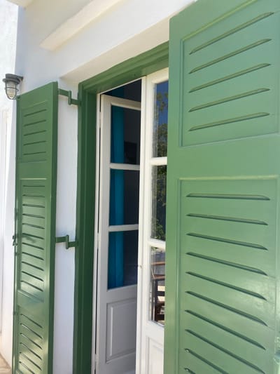 The scene captures an exterior architectural detail of a building in Πλατύς Γιαλός, Greece, under bright daylight. A white-painted double window or door, featuring multiple glass panes and white mullions, is partially open. Two louvered shutters, painted a light green, are fully opened and secured against the white-washed exterior wall by dark metal latches. A dark-colored, caged nautical-style wall lamp is mounted on the white wall to the left of the opening. Reflections of blue sky and green foliage are visible on the window glass. Through the partially open window, a room interior featuring a distinct blue wall or curtain is discernible. No individuals or active interactions are present within the frame.
