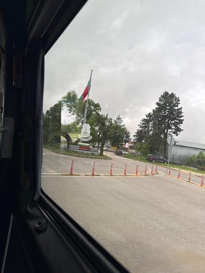 View from inside a vehicle looking through an open sunroof. The scene outside is a road leading to Brestovets, Bulgaria, as indicated by a sign with Bulgarian writing and a Bulgarian flag. The sky is overcast. There is a road with orange and white traffic cones on the right side, and a building in the background. Some trees are also visible.