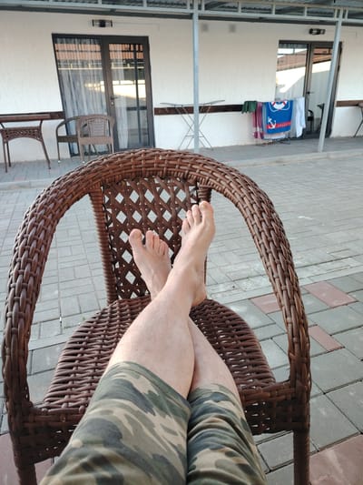 A person's bare feet, crossed at the ankles, are resting on a wicker chair. The person is wearing camouflage shorts. The chair is situated on a paved patio area with a tiled surface. In the background, there is a white building with doors and windows, and a clothesline with laundry hanging on it. The scene appears to be outdoors, possibly in a residential or hotel area. The location is identified as Toranghylyq, Kazakhstan.