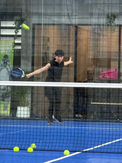 The image captures a young man playing padel on an indoor court in Ban Buak Khrok North, Thailand. The court is vibrant blue with white markings and a black net divides the playing area. The man, dressed in a black sleeveless hoodie and black athletic pants, is in mid-action, swinging a padel racket at a bright yellow ball in the air. His body is positioned to make a powerful stroke, and his gaze is focused on the ball.
The background reveals the indoor setting, with large glass panels reflecting the environment. Through the glass, we can see a potted plant with lush green leaves, a yellow poster with text that reads "LEVEL UP YOUR SKILLS WITH NIKKO PADEL," and some indistinct figures in the distance. A bright pink sports bag is also visible in the background. On the court, several yellow padel balls are scattered on the blue surface, some near the net. The lighting appears to be artificial, creating a somewhat diffused and reflective atmosphere within the court. The overall scene depicts an active sports moment within a modern indoor recreational facility.