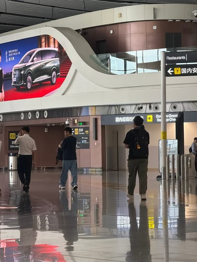 This image depicts an indoor scene, likely an airport or large public building, with people walking in a brightly lit hallway. The floor is highly reflective, showing distorted images of the ceiling lights and people. In the background, there are signs with Chinese and English text, indicating directions to "Restaurants" and "Domestic Overs..." suggesting a transportation hub. One sign points to "Elevator". A large digital billboard displays an advertisement for a car, a dark green minivan. Several individuals are walking away from the camera, dressed in casual attire. One person is wearing a black backpack, and another is carrying a bag over their shoulder. The architecture suggests a modern and spacious environment, with smooth, curved surfaces and metallic elements. The lighting is overhead and appears to be a mix of natural and artificial sources, creating a bright, clean atmosphere. There is no clear indication of the specific city, but the presence of Chinese signage implies a location within China. The overall impression is one of transit and modern public space.