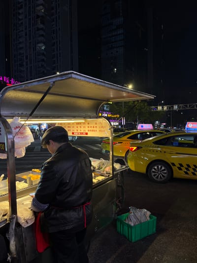 A person stands at a food cart with a menu written in Chinese. The menu lists prices for various items, including "豆成 (soy milk) 2 yuan," "麻园 (sesame cake) 2 yuan," "油条 (fried dough stick) 2 yuan," and "馒头 (steamed bun) 1 yuan." A yellow taxi cab is parked nearby, and a green crate sits next to the cart. The scene is illuminated by streetlights and the cart's own lighting, set against a backdrop of buildings at night.