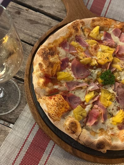 The image displays a prepared pizza on a round wooden serving board with a dark rim, positioned on a rustic wooden plank table in a dining setting within Thành phố Phan Thiết, Vietnam. The pizza, centrally located, features a golden-brown, slightly charred crust. Its toppings include melted white cheese, numerous irregularly shaped yellow pineapple chunks, and slices of light pink ham. A small sprig of fresh green parsley garnishes the pizza towards the center-right. To the upper left, a clear stemmed glass, partially filled with a pale yellow liquid and displaying condensation, is visible. A placemat with white and red stripes is partially visible beneath the wooden board and glass in the lower-left section of the frame. No individuals are present in the scene.
