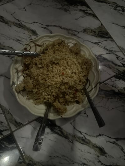 A close-up overhead shot shows a white, scalloped-edge plate filled with a mound of cooked rice on a marble-patterned tiled surface. Two silver forks are present, one resting on the left edge of the plate and partially submerged in the rice, and another lying on the right side of the plate. The lighting appears dim. The location is the city of Pantinapu, Nigeria.