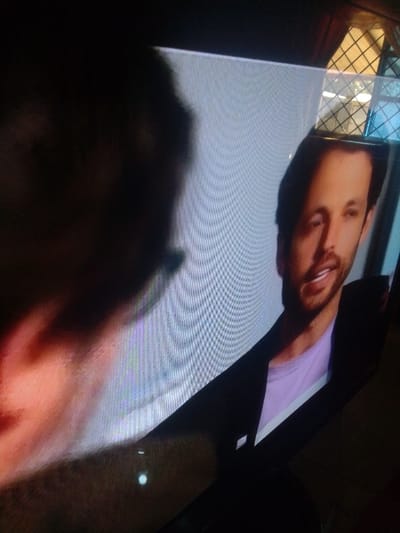The image displays a television screen presenting a still or video frame. On the screen's right side, a light-skinned male subject with short dark hair and a beard or stubble is visible from the chest up, oriented slightly left. He wears a dark jacket over a light purple t-shirt, with his mouth slightly ajar. A distinct vertical shadow divides his face. The screen's left portion shows a dark, blurry, amorphous shape, potentially a reflection or out-of-focus silhouette, with a reddish hue at its lower edge. The screen itself exhibits prominent horizontal and vertical pixel lines. Reflections are present on the screen: in the upper right, a window with a dark grid pattern is reflected, and near the bottom center, a small, bright, circular light source is visible. The television is encased in a dark bezel. The physical location of the recording, Mista Ali, Nigeria, cannot be determined from the visual information provided within the image.