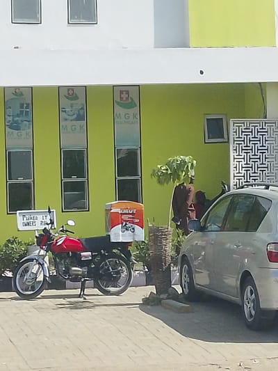 This image captures an outdoor scene in Darmanawa, Nigeria, depicting the facade of a commercial building and a section of its forecourt. The building's lower level is painted a vibrant light green, topped by a white upper section, both featuring dark-paned rectangular windows. Three vertical banners are affixed to the green wall, each displaying a red cross on a white shield logo and the text "MGK." The banners identify the establishment as "MGK HEALTHCARE," "MGK DIAGNOSTIC CENTRE," and "MGK CLINICAL LABS" (partially visible). To the right of the banners, a decorative white lattice screen is present.
In the paved forecourt, a red motorcycle with silver elements is parked on a kickstand on light-colored interlocking pavers. The motorcycle features a large, orange and white delivery box, marked "OMUT" with a motorcycle graphic, affixed to its rear rack. Adjacent to the motorcycle, a silver four-door sedan is parked, its rear passenger side and bumper visible. A white rectangular sign with black text reading "PARKED AT OWNERS RISK" is mounted on a pole positioned near the front wheel of the motorcycle.
Green foliage lines the base of the building, and a small tree with sparse green leaves stands between the motorcycle and the sedan. A woven cylindrical planter is situated near the tree. Partially obscured by the tree and the sedan, a dark-clad figure, appearing to be a mannequin or statue, stands upright.