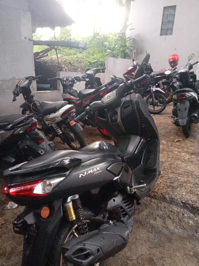 The image shows a row of motorcycles parked in front of a white building in Kecamatan Caringin, Indonesia, located at latitude -7.523 and longitude 107.485. The motorcycles are parked in a row, with the closest one to the viewer being a black Yamaha NMax with a gold inscription on the side. Behind it are several other motorcycles, some of which are red, silver, and black. All the motorcycles are parked in front of a white building, with a window on the side. The building is surrounded by greenery, including trees and bushes. The scene appears to be shot from the perspective of someone standing in front of the motorcycles, looking towards the building. The ground is wet and muddy, suggesting recent rainfall. The scene is devoid of people.