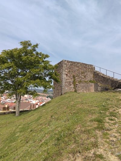 A stone structure sits on a grassy hill overlooking a town, likely in Mogadouro, Portugal. The structure appears to be part of an old fortress or castle, with an external stone staircase and metal railing visible. A tree stands near the structure. The hill slopes down towards the town, which consists of buildings with red-tiled roofs. The sky is partly cloudy with a light blue hue. The overall impression is a historical site with a scenic view. No people are visible in the image.