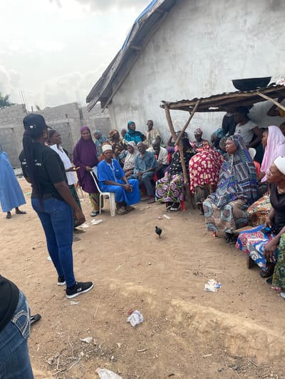 An outdoor community gathering is depicted in Osara Gada, Nigeria. The scene features approximately 25-30 individuals, predominantly adults, congregated on a dirt ground area. To the right, a weathered concrete building with a corrugated metal roof is visible, with a large black basin resting on a wooden support structure against its wall. Adjacent to this building is a rudimentary wooden shelter. To the left, uncompleted cinder block construction is present.
In the foreground, a woman with dark skin, wearing a black t-shirt with partial text ("...SION," "Y," "COMMUNITY," "EXCELLENCE"), blue jeans, and black sneakers, stands with her back to the viewer, facing the assembled group. She gestures with her right hand, suggesting she is addressing the attendees.
The majority of the group is seated on various plastic chairs and low wooden benches or stools, forming a semi-circle. Many individuals wear traditional Nigerian attire, characterized by colorful patterned fabrics and head coverings, particularly among the women. A male individual in the center is seated on a white plastic chair, wearing a blue traditional garment and a cap, holding a walking stick.
The ground is unpaved dirt, littered with discarded items including plastic bottles, wrappers, and other debris. A small, black chicken walks on the ground in the mid-ground, moving away from the assembled group. The sky overhead is overcast.