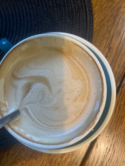 A close-up, overhead shot focuses on a creamy, frothy beverage in a ceramic mug, likely coffee or latte, with a spoon resting on the rim. The drink exhibits latte art, with a swirl pattern formed by the foam and a darker liquid. The mug, with its white base and teal and brown stripes, sits on a wooden surface. To the left, a dark, textured placemat is partially visible, and behind it, a teal ceramic handle of the mug. The scene is set in Berehove, Ukraine.