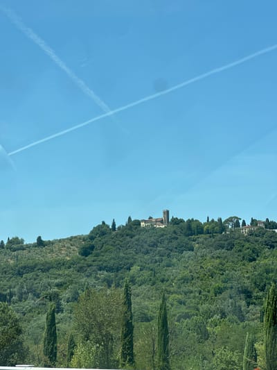 The image shows a clear blue sky with two contrails crisscrossing. Below the sky, a lush, green hillside is visible, densely covered with trees and vegetation. Atop the hill, there is a small cluster of buildings, including a prominent tower with a church or monastery adjacent to it, characteristic of the Tuscan landscape. The buildings appear to be made of stone and have red-tiled roofs. The overall scene suggests a tranquil, rural setting. This view is likely from the vicinity of Bagno a Ripoli, Italy, given the characteristic rolling hills and architecture of the region.