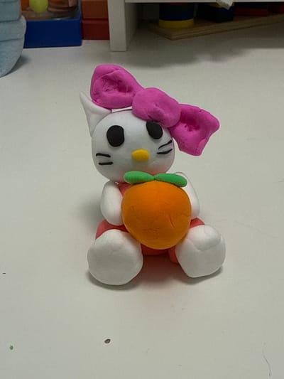 The image displays a close-up, horizontal shot of a handmade clay figurine, likely intended to resemble Hello Kitty, positioned against a plain white wall. The figurine is predominantly white with a large pink bow on its head and black oval eyes and whiskers. A yellow dot on its forehead may represent a nose. It is embracing a bright orange sphere, possibly an apple or orange, adorned with a small green stem. The figurine has white, rounded limbs.

To the left of the figurine, a shelf unit is partially visible. This unit contains colorful building blocks, including red, blue, and yellow ones, suggesting a child's play area or toy storage. The lighting appears to be artificial, casting soft shadows and highlighting the texture of the clay. The overall scene suggests a domestic setting, possibly a child's room, in Beryozovsky, Russia. There is no discernible time of day, weather, or specific event taking place beyond the display of the handmade toy. No visible text is present in the image.